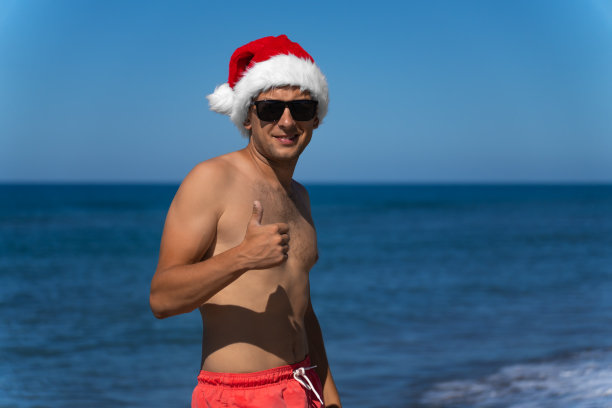 年轻英俊的男子戴着太阳镜，穿着珊瑚色的游泳短裤，戴着圣诞老人帽，在海滩上竖起大拇指，复制空间。圣诞假期图片下载