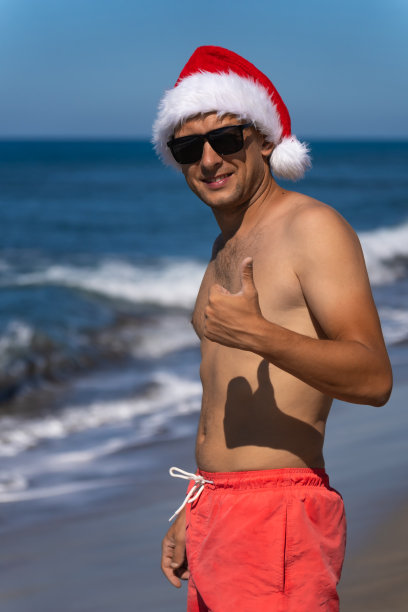 一个面带微笑的英俊年轻人，戴着太阳镜，穿着珊瑚色的游泳短裤，戴着圣诞老人的帽子，在海边伸出手指。圣诞假期。垂直图图片下载