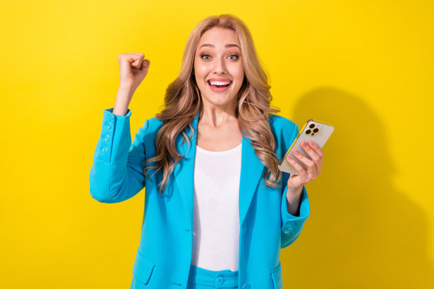欣喜的美女手持智能手机举起拳头成功成就的照片被隔离在黄色背景上图片下载