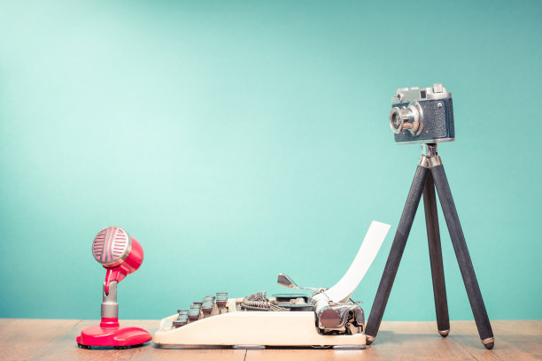 20世纪50年代的老式麦克风，三脚架上的过时胶片相机和木制办公桌前薄荷蓝色墙壁背景上的旧打字机。博客的概念。复古滤镜照片图片下载