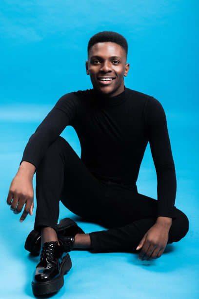 全身照片快乐的微笑的非洲裔美国男人模特坐在地板上的蓝色背景的工作室孤立的摆出不同的姿势穿着时尚的黑色毛衣和牛仔裤图片下载