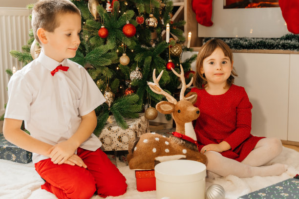 哥哥和妹妹坐在圣诞树旁边，享受着礼物。圣诞图片，树和灯的背景。孩子们在圣诞树旁打开新年礼物。圣诞快乐，新年快乐。图片下载