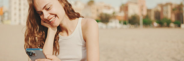 在现代建筑背景的沙滩上，微笑的女孩手里拿着手机坐在沙滩上。漂亮的女孩正在用她的智能手机看视频和照片图片下载