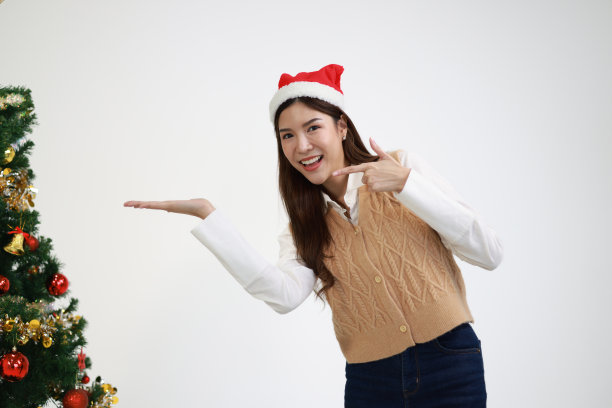 一位戴着圣诞帽的快乐女子用手指着圣诞树，这是圣诞节的概念图片下载