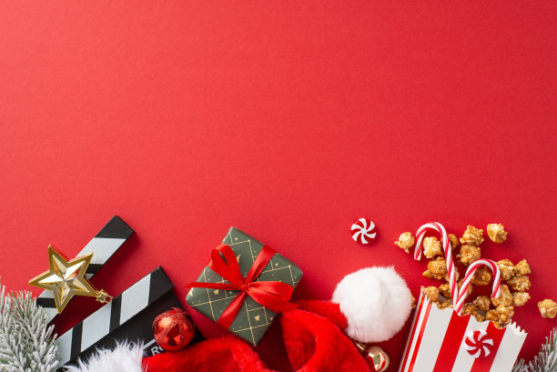 圣诞电影之夜，爆米花点子。电影拍摄者的头顶照片，美味的爆米花在条纹盒，圣诞老人的帽子，小装饰品，星星装饰，糖果，霜冷杉树枝在红色背景与广告空间图片下载