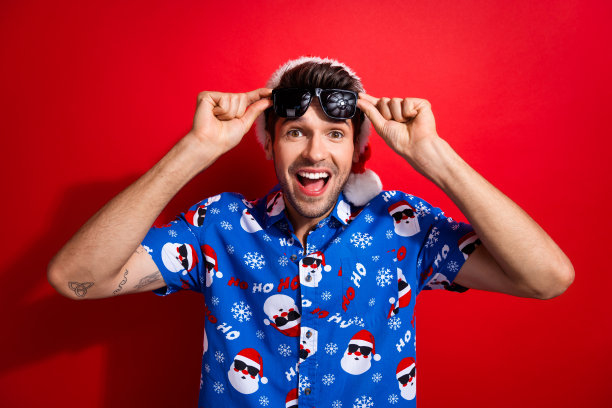 照片肖像英俊的年轻人兴奋触摸太阳镜戴圣诞老人印刷圣诞服装孤立的红色背景图片下载