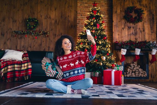 迷人的黑皮肤女孩坐在地毯上聊天视频通话的照片，显示了房间里的盒子，神奇的气氛照亮了树图片下载