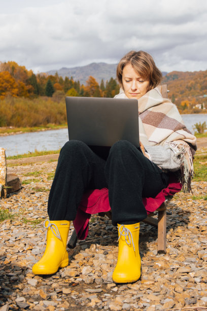 拿着笔记本电脑的女孩站在美丽的山景上。在喀尔巴阡山脉河岸工作的自由职业者。秋山田园诗般的职场。自由的概念。远程工作。图片下载
