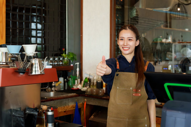 漂亮的亚洲女商人穿着工装裤站在柜台的咖啡机后面，她微笑着竖起大拇指。拍照片宣传一家小咖啡店图片下载
