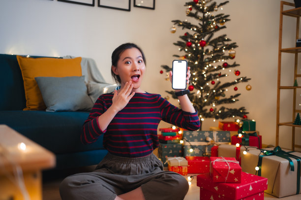 一名亚洲女性坐在装饰着圣诞树的客厅里，拿着白色屏幕的智能手机，开心而兴奋图片下载