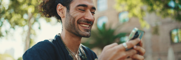 马尾辫和胡茬的意大利帅哥坐在老建筑背景的长椅上玩手机。时尚男士在智能手机上写信息、看照片图片下载