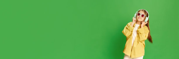 宽照片小女孩穿着时髦，听音乐在绿色背景与微笑看着相机。科技、代际、童年、生活方式、广告的概念。图片下载