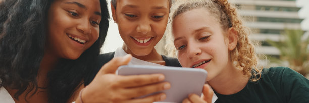 三个十几岁前的女孩朋友正坐在海边玩手机，看视频、照片。三个青少年在户外的城市景观背景图片下载