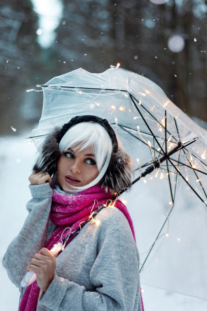 女人，戴着白色假发，戴着保暖耳罩，穿着舒适的毛衣，围着粉红色的围巾。她拿着一把透明的伞，上面镶嵌着闪烁的灯光，营造出一种神奇的冬季氛围图片下载