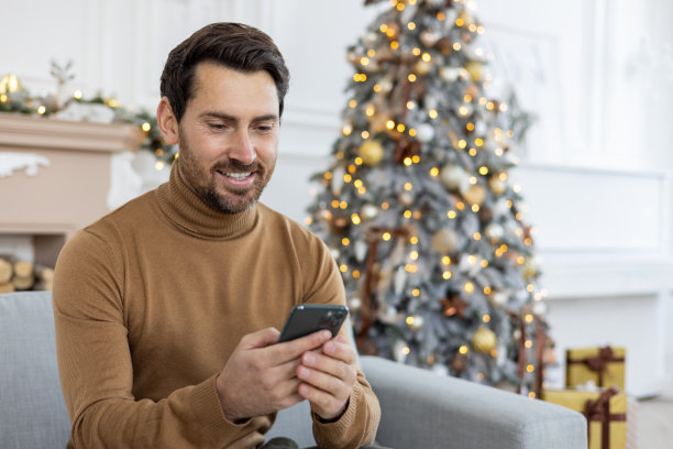 新年假期和周末，一个面带微笑的年轻人坐在家里圣诞树旁边的沙发上玩手机。特写照片图片下载