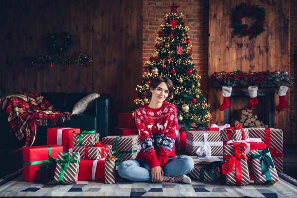 照片中可爱的年轻女士盘腿坐在地板上，赠送一堆穿着时髦的红毛衣，装饰着圣诞树的室内客厅图片下载