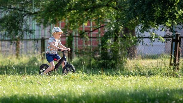 一个小男孩戴着帽子，穿着蓝色短裤，在一个炎热的夏天骑着自行车在一个城市公园，横向侧面照片。图片下载