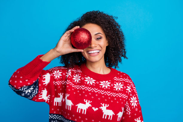 照片的快乐可爱的女孩穿着时髦的装饰品套头衫，手拿红色闪亮的玩具冬青快乐圣诞精神孤立的蓝色背景图片下载