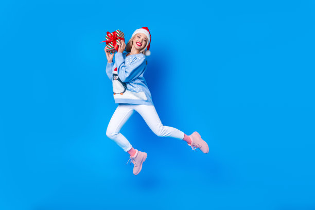 全身照片跳跃活跃的年轻女子红色圣诞老人头饰，企鹅印花衬衫保持惊喜孤立的蓝色背景图片下载