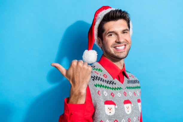 照片中可爱的年轻人指着空旷的空间露出牙齿微笑，穿着圣诞印花背心，帽子，衣服孤立在蓝色背景上图片下载