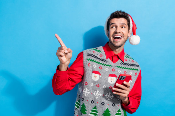 照片肖像可爱的年轻人看点空的空间拿着装置穿圣诞印花背心帽子衣服孤立在蓝色的背景图片下载