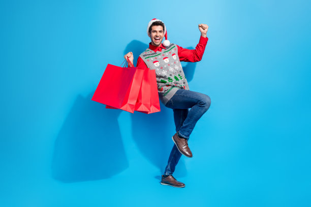全身照片欣喜若狂的家伙戴着圣诞帽，拿着新衣服庆祝节日孤立的蓝色背景图片下载
