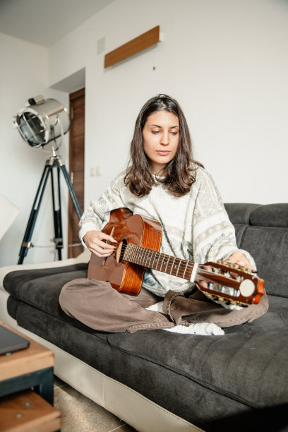 年轻女子坐在沙发上，看着她的笔记本电脑，并调整吉他-库存照片图片下载