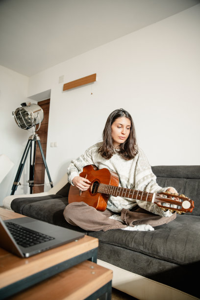 年轻女子坐在沙发上，看着她的笔记本电脑，并调整吉他-库存照片图片下载