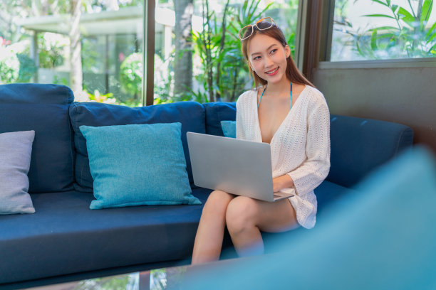 穿着沙滩装，戴着墨镜的年轻亚洲女子在热带咖啡馆里用笔记本电脑工作。女性数字游民在度假氛围中用笔记本电脑工作。穿着沙滩装的亚洲女人坐在咖啡馆的沙发上用笔记本电脑远程工作。图片下载