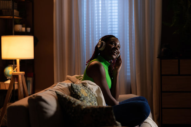 远处的照片漂亮的黑皮肤的美国黑人妇女戴着新的现代耳机微笑梦想抬头，而坐在家里的沙发上的莲花姿势享受时刻图片下载