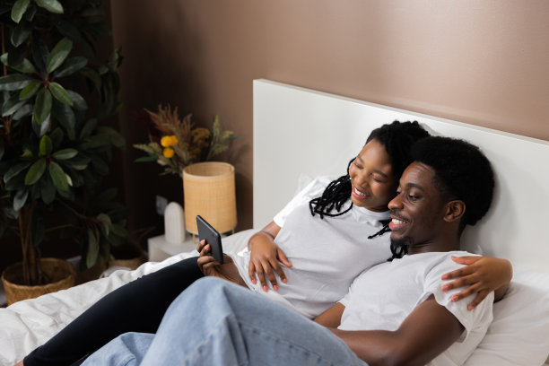 幸福家庭概念。深色皮肤的非洲裔男女躺在床上，穿着休闲的白色t恤，拿着手机在社交网络上查看新闻，看朋友的照片和视频图片下载