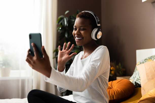 照片俯视图兴奋的非洲裔美国人，皮肤黝黑，手里拿着手机，戴着新的现代耳机，通过视频链接说话，同时在家电脑上在线工作图片下载