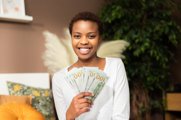 美国黑人年轻女子穿着休闲装坐在家里的床上数钱的照片。看看我有多少钱，快乐概念钞图片下载