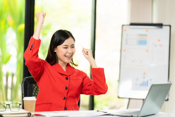 快乐的年轻亚洲女性在办公室的笔记本电脑前高举双臂庆祝成功，金融商业概念图片下载