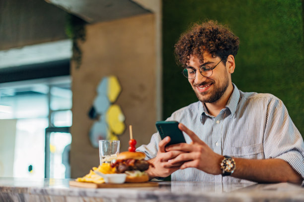 在开始吃美味的汉堡之前，一个面带微笑的男人坐在餐厅里玩手机图片下载
