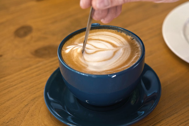 手正在搅拌拿铁的奶泡。用钢勺搅拌卡布奇诺。热咖啡在白色经典陶瓷杯在木桌。现代咖啡店，自助餐厅。厚的泡沫。特写镜头图片下载