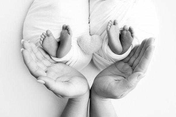 爸爸和妈妈抱着刚出生的双胞胎的脚。父母的手和孩子的脚在包裹和编织的心。新生双胞胎的腿、脚趾、脚和脚跟。黑白照片。图片下载