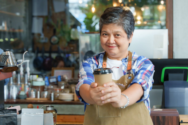 白发的退休亚洲女商人站在前面拿着一次性的棕色咖啡杯微笑。站在咖啡机柜台前为小型家庭咖啡馆拍摄宣传照片。图片下载