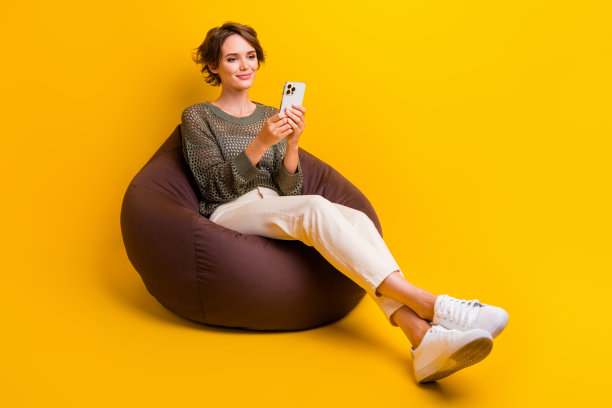 照片中可爱可爱的女孩穿着时尚的衣服，坐在舒适的沙发上写短信，空白的空间孤立在黄色的背景上图片下载