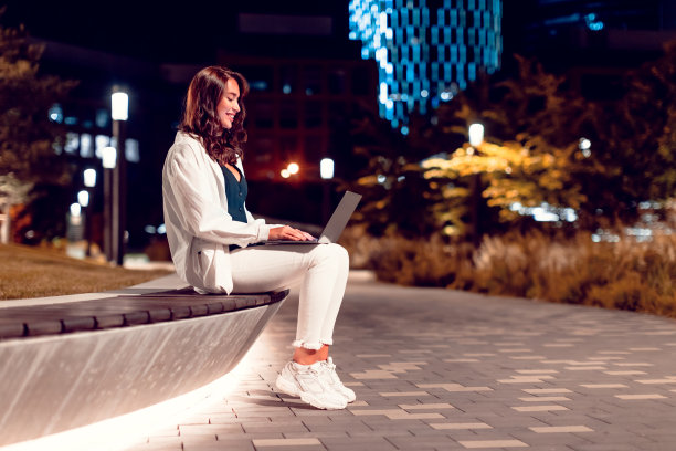 快乐的自由职业者女士在笔记本电脑上工作，坐在晚上公园的长凳上在户外工作，侧视图，自由空间图片下载