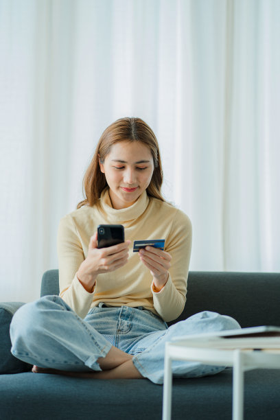 开朗的亚洲女性使用智能手机，网上购物，手持信用卡，支付，坐在家里的沙发上网上银行和电子商务应用垂直图片图片下载
