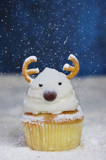 个人特写图片，自制的圣诞驯鹿设计纸杯蛋糕，上面撒着筛过的糖粉雪，奶油漩涡顶部装饰着糖果包裹的巧克力甜鼻子和盐椒盐脆饼鹿角，斑纹蓝色背景，重点在前景图片下载