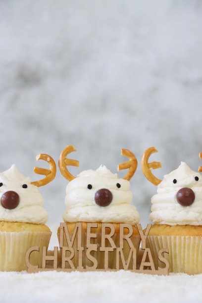 圣诞快乐标志的特写图片，对着三个自制的圣诞驯鹿设计纸杯蛋糕，奶油漩涡顶部装饰着糖果涂层的巧克力甜鼻子和盐椒盐卷饼鹿角，糖雪，重点在前景，复制空间图片下载