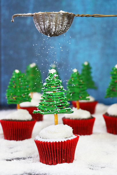 图片中，糖霜筛过一批自制的，圣诞树设计的巧克力纸杯蛋糕，奶油涂在干燥的椰子顶部，白巧克力和椒盐脆饼棒树，展示在白雪皑皑的针叶林场景，蓝色背景图片下载