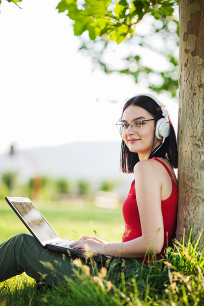 深棕色头发的女孩戴着白色耳机在公园的树下使用笔记本电脑图片下载
