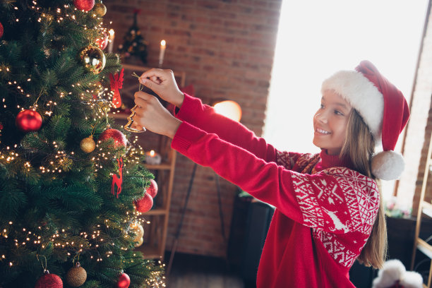 照片中，开朗积极的小女士穿着红色印花套头衫，在家里的房间里享受挂圣诞圣诞树装饰图片下载