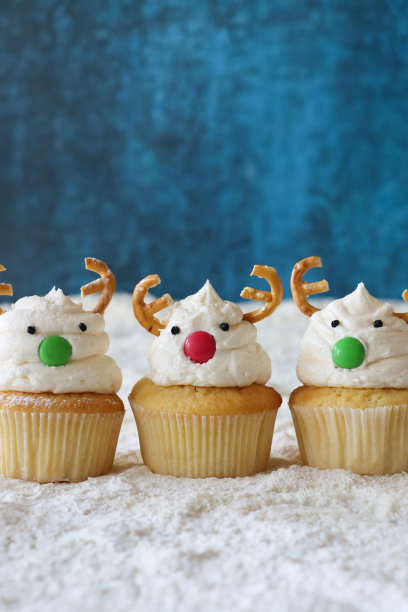 特写图片一排三个，自制的，圣诞驯鹿设计的纸杯蛋糕，奶油漩涡顶部装饰有糖果包裹的巧克力甜鼻子和咸椒盐卷饼鹿角，糖雪，斑纹蓝色背景，重点在前景，复制空间图片下载
