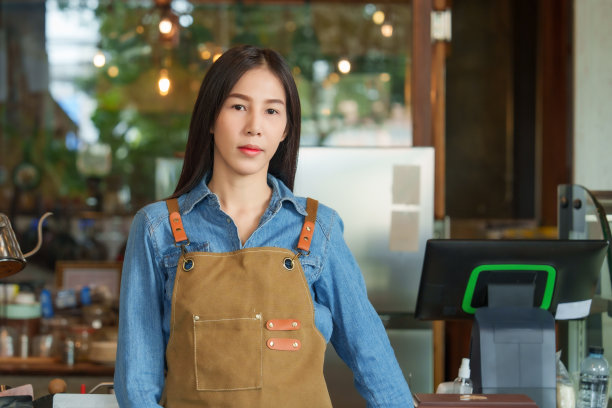 年轻的亚洲女子海军蓝长袖连衣裙戴着可爱的棕色围裙年轻的女商人咖啡馆，手持平板电脑面向前拍宣传照片咖啡馆的前台柜台。咖啡馆，小型家族企业。图片下载