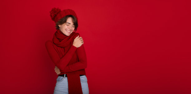 大横幅照片开朗的年轻女子提出季节性冬季促销优惠在红色复制空间。可爱的黑发女士在红色冬季围巾，毛衣和帽子。新年和圣诞节图片下载