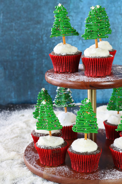 一批自制的，圣诞树设计的巧克力纸杯蛋糕的特写图片，蛋糕架上的奶油涂在干燥的椰子顶部，白巧克力和椒盐脆饼棒树，糖粉雪，斑驳的蓝色背景，复制空间图片下载